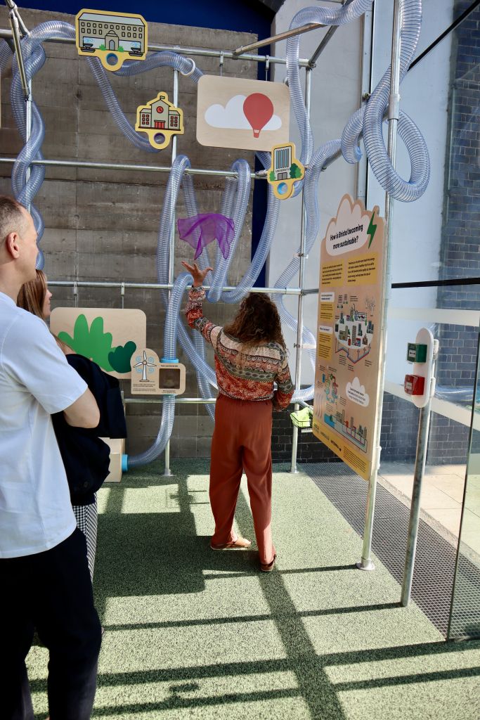 Visitor participating in an interactive activity by tossing a pink fabric piece into a network of transparent tubes within a sustainability-themed exhibition display.