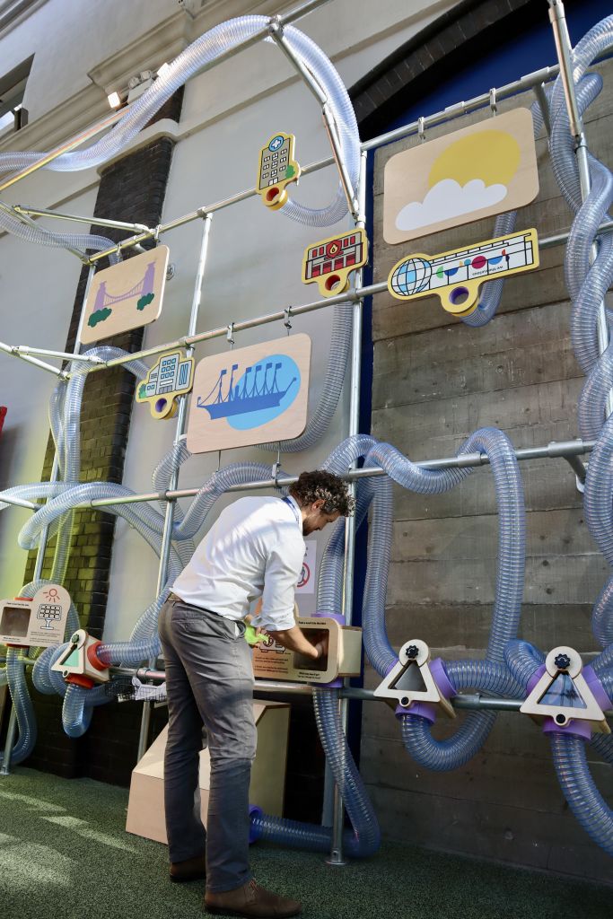 Visitor using a hands-on exhibit component connected to a network of transparent tubes within an educational display about sustainability and renewable energy.