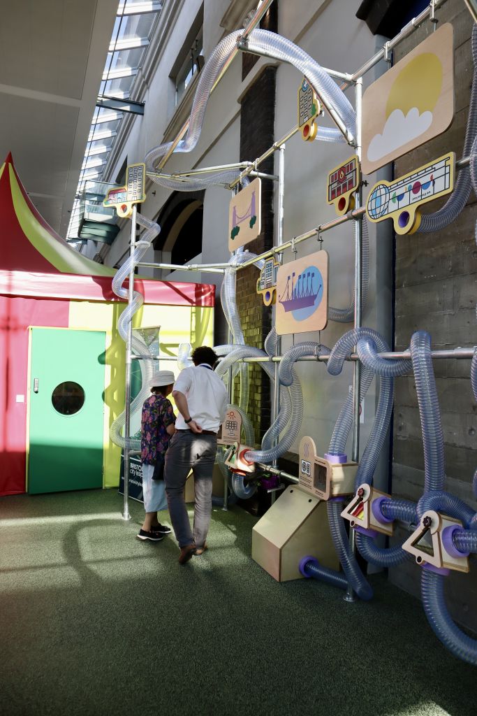 Wide view of an interactive sustainability installation featuring transparent tubing, illustrated Bristol landmarks and educational displays inside a contemporary exhibition space.