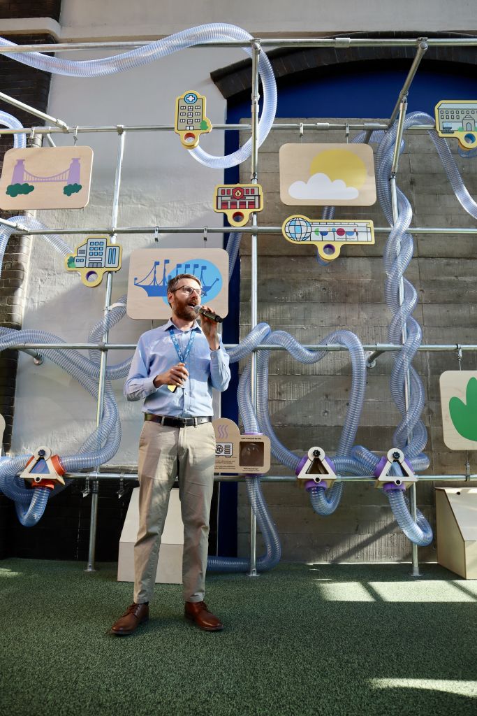 Speaker using a microphone to present beside an interactive exhibition about sustainability, with transparent tubing and illustrated city-themed displays forming the backdrop.
