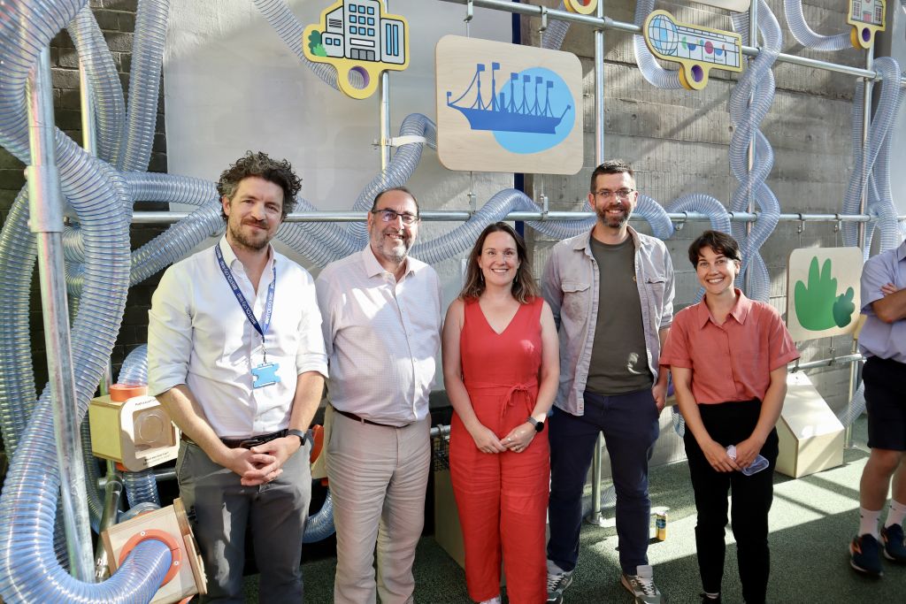 Five attendees standing in front of an interactive sustainability-themed installation made of transparent tubes, illustrated city icons and educational displays inside a public exhibition space.