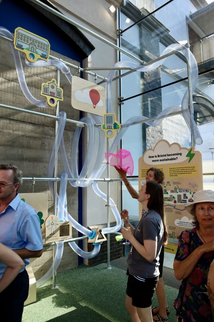 Visitors explore an interactive exhibit featuring a network of transparent tubes and illustrated Bristol landmarks, with one visitor using a pink fabric piece as part of a hands-on activity.