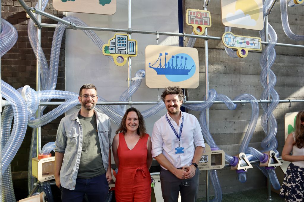Three attendees posing in front of an interactive exhibition about sustainable energy in Bristol, surrounded by transparent tubing and illustrated city-themed display panels.