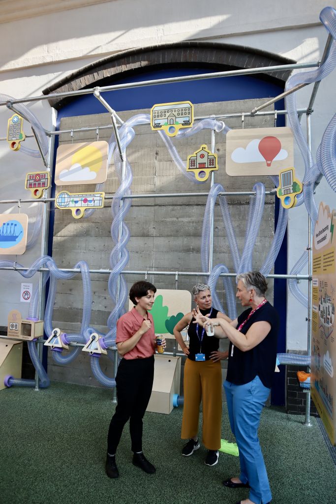 Three attendees in conversation beside an interactive installation about Bristol’s sustainable future, featuring interconnected transparent tubes and illustrated city icons.