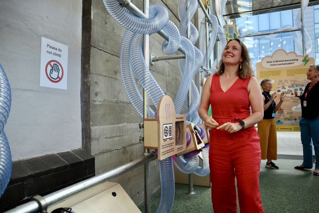 Visitor standing beside an interactive exhibit, looking up at a network of transparent tubes and educational sustainability displays, with an information board visible in the background.