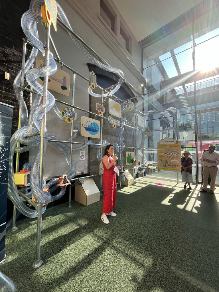 Speaker addressing guests at the launch of an interactive sustainability exhibition, standing beside a large installation featuring transparent tubes and Bristol-themed illustrations.