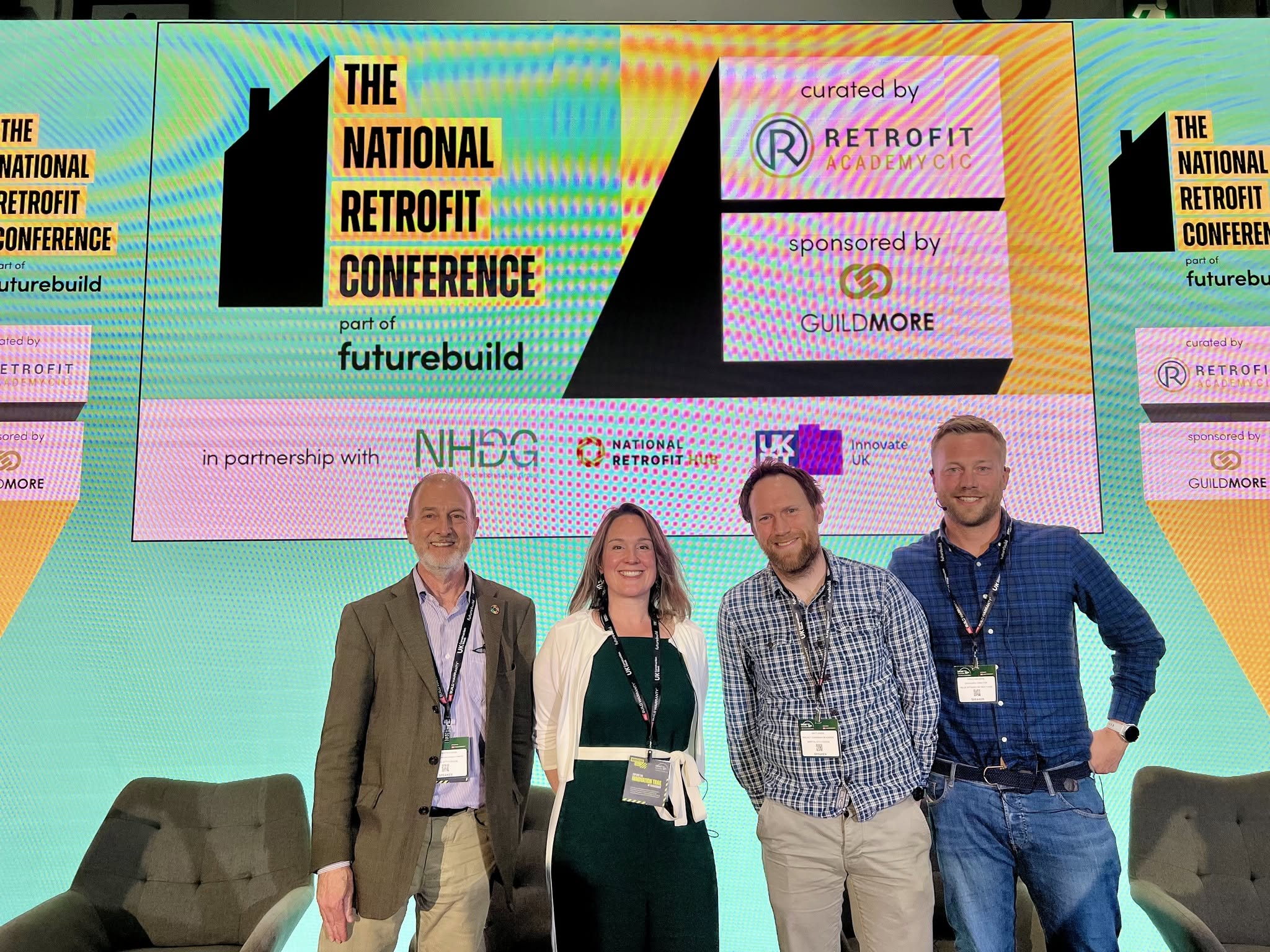 Four panel speakers stand on a conference stage beneath a large screen displaying branding for The National Retrofit Conference, part of Futurebuild. The screen features event partner and sponsor logos, including Retrofit Academy CIC and Guildmore. Two armchairs are positioned on either side of the stage. The image was taken during a panel discussion on retrofit, energy efficiency and decarbonisation in Bristol and the South West.
