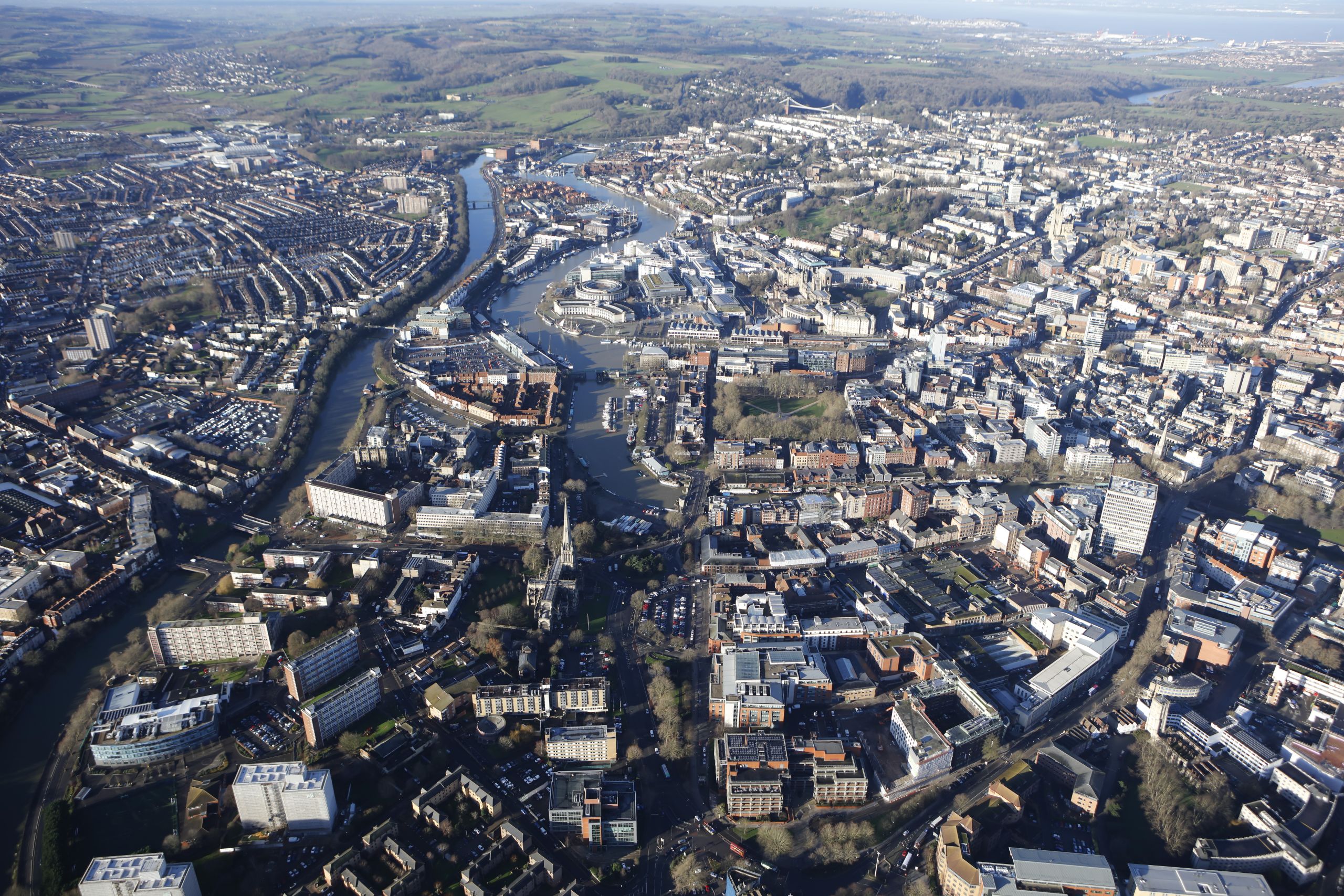 An aerial view of central Bristol showing the River Avon winding through the city, with harbourside developments, residential neighbourhoods and surrounding green countryside.